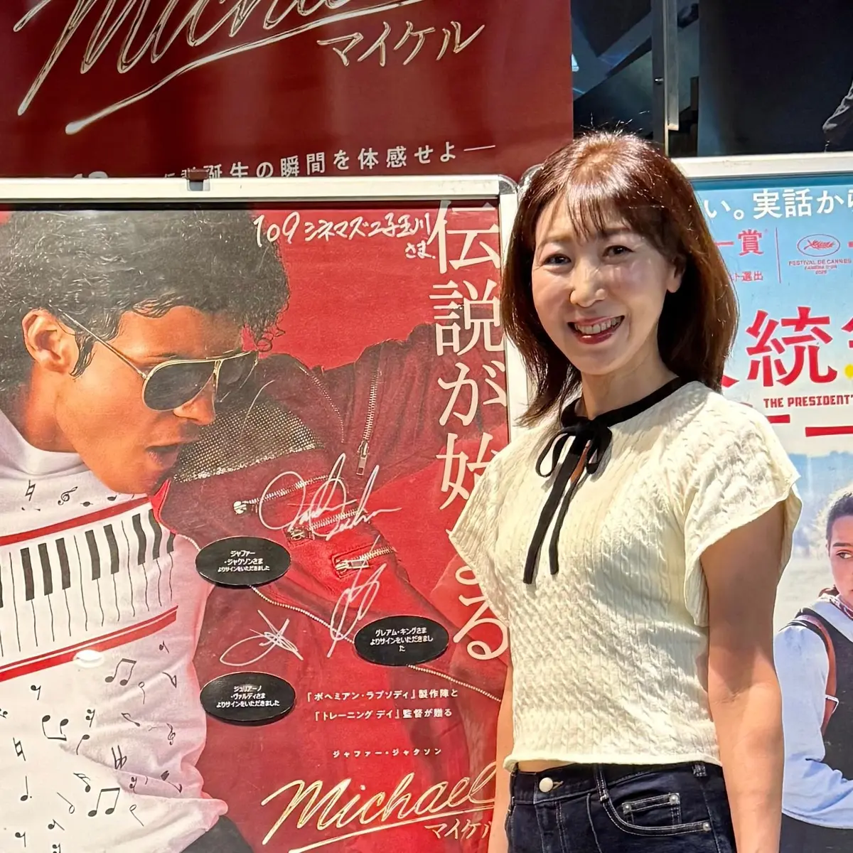 マイケル・ジャクソン展のポスターの前で、女性が笑顔で記念撮影をしている。ポスターにはマイケル・ジャクソンの写真と、日本語のテキストが記載されている。背景には他の映画ポスターも見られる。