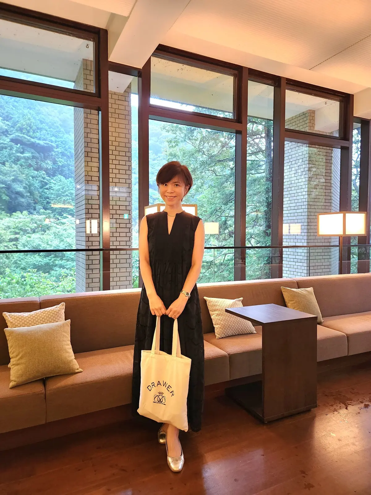 黒いワンピースを着た女性が、窓際に置かれたソファの前に立っている。背景には緑豊かな自然が広がり、窓枠は木目調のデザイン。右手に「Drawer」のロゴが入った白いトートバッグを持ち、シルバーの靴を履いている。