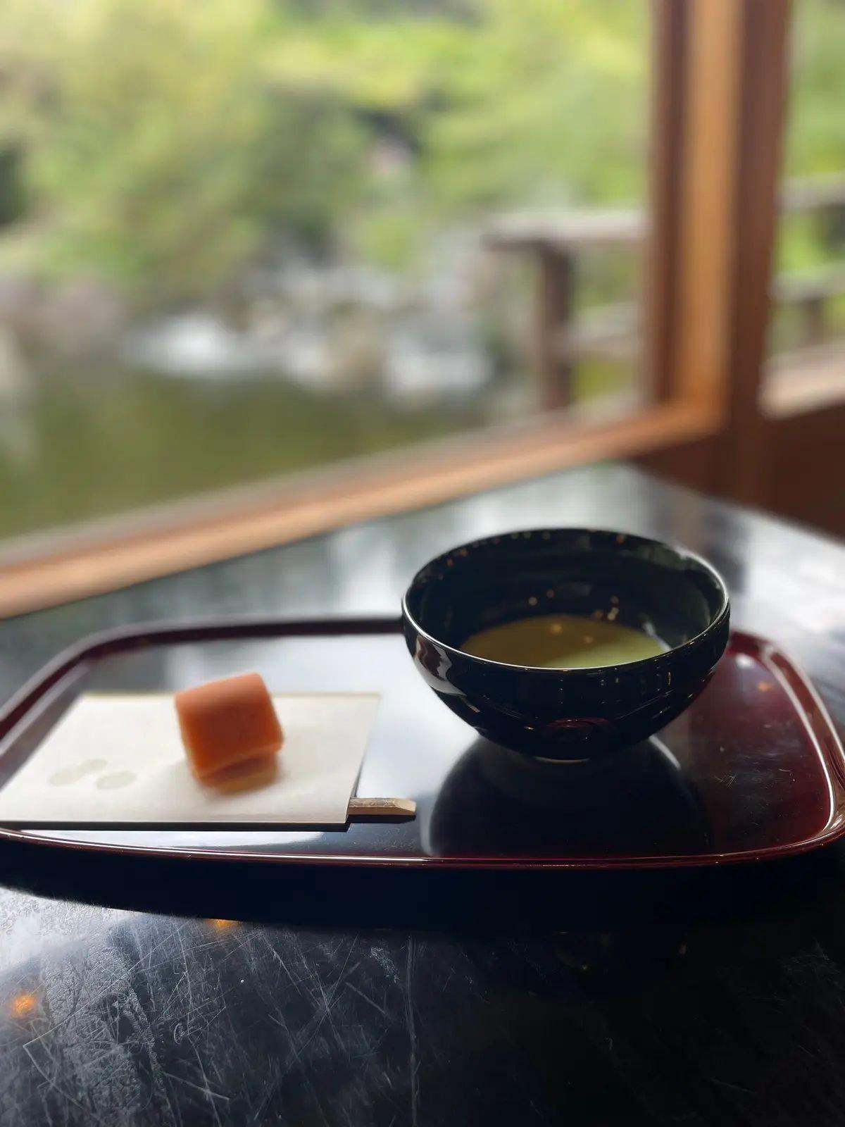 漆塗りの長方形の盆に、和菓子と抹茶が置かれている。和菓子は薄いピンク色で、抹茶は黒い茶碗に入っている。背景には窓から見える緑豊かな庭園。