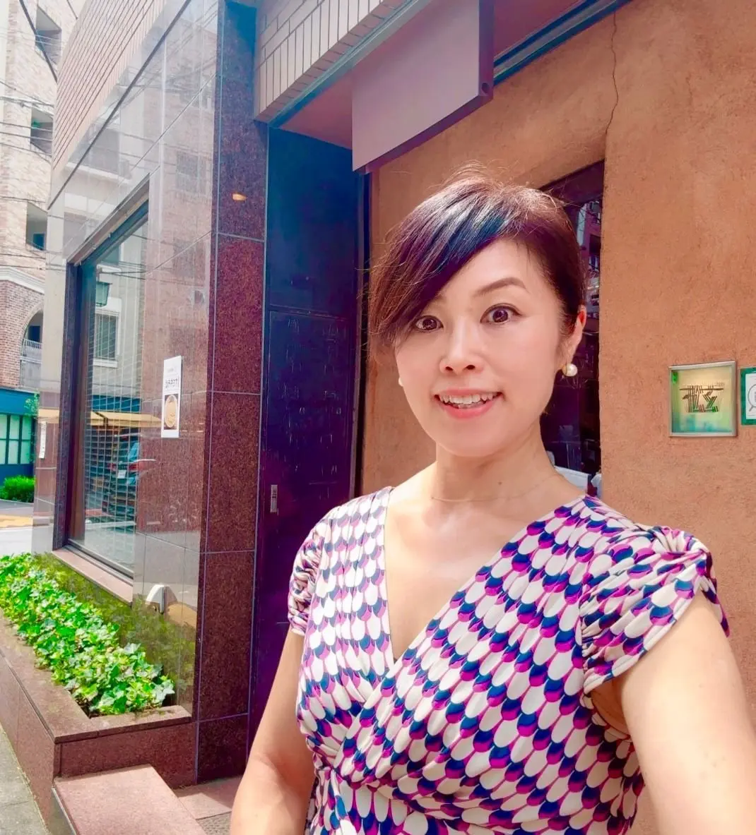 笑顔の女性が、建物の入り口付近でこちらを見ている。紫、白、黒の幾何学模様のワンピースを着ており、背景には高層ビルと緑の植栽が見える。