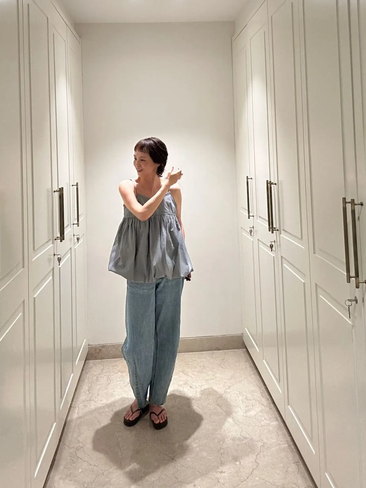 グレーのサロペットとサンダルを履いた女性が、白い壁の廊下に立ち、片手を頭に当てて微笑んでいる