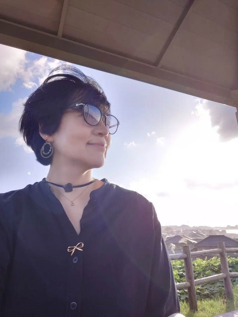 黒髪の女性が、屋外の木造の屋根の下で横顔を見ている。黒縁の眼鏡をかけ、黒いワンピースを着ている。首には緑色のチョーカーと金色のネックレスを着用し、耳にはリング型のピアスをしている。背景には青空と白い雲、そして遠くの風景が見える。