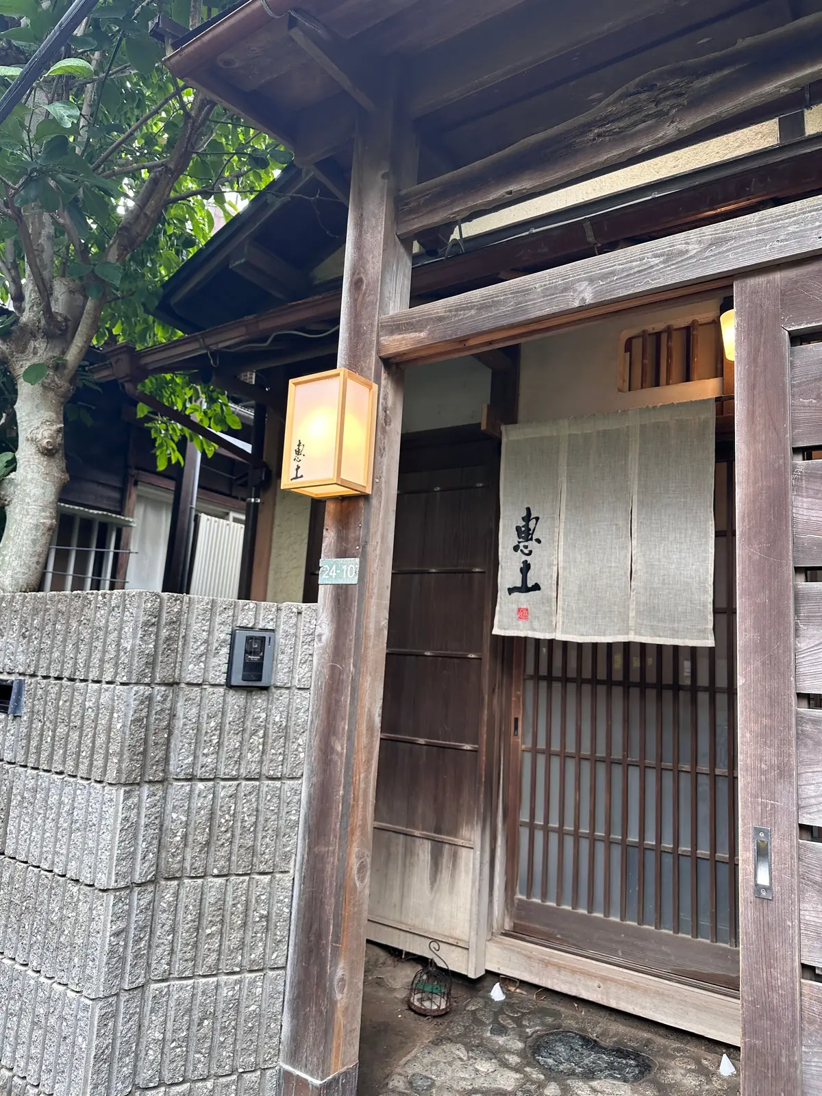 木造の建物と石積みの塀。建物の入り口には提灯と、書かれた白い掛け軸が飾られている。住所表示のプレートも見える。