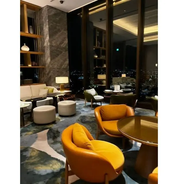 夜景を望む高級ホテルの一室。オレンジ色の椅子と丸テーブルが配置され、白いプフやソファ、緑色の椅子もある。抽象的な模様のカーペットが敷かれ、壁には棚と装飾品が置かれている。