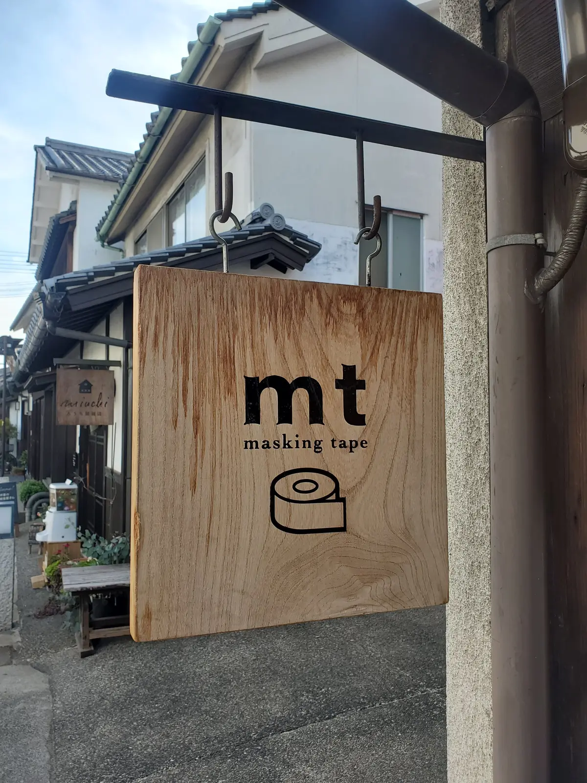 木製の長方形の看板に、黒字で「mt masking tape」と記載。テープのイラストが右下に配置され、背景には日本の古い街並みが見える。