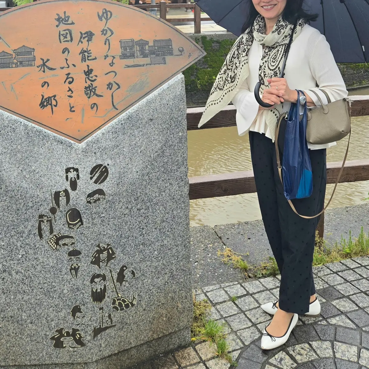 女性が石碑の横に立ち、傘を手に持つ。石碑には日本語の文字とイラストが描かれている。背景には水路と木製の橋が見える。