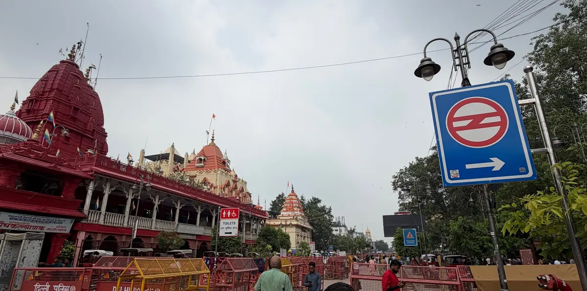 インドのデリーにある赤色の寺院と、その周辺の道路の様子。歩行者が数人おり、右側には地下鉄の標識と警察のバリケードが見える。空は曇り。