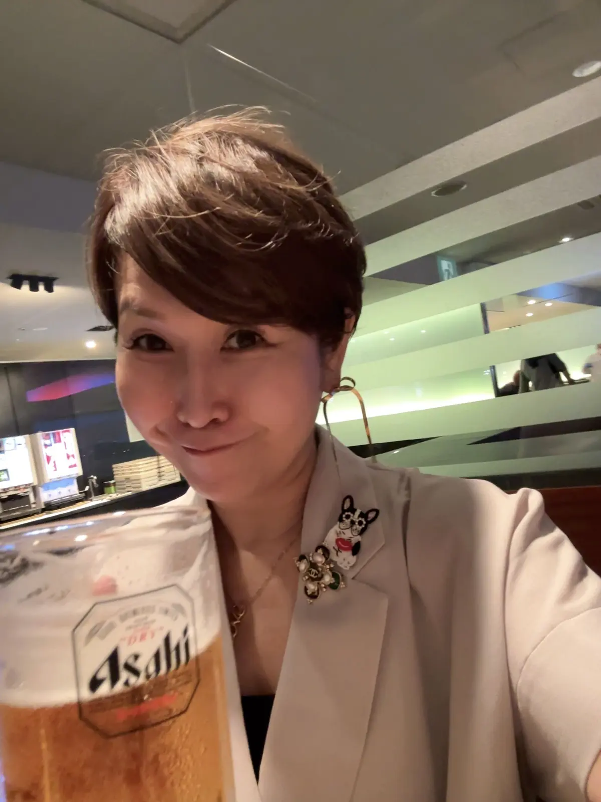 笑顔の女性が、アサヒスーパードライのビールを手にしているセルフィー。白いジャケットを着用し、胸元に犬のピンバッジをつけている。背景はレストランのような雰囲気。