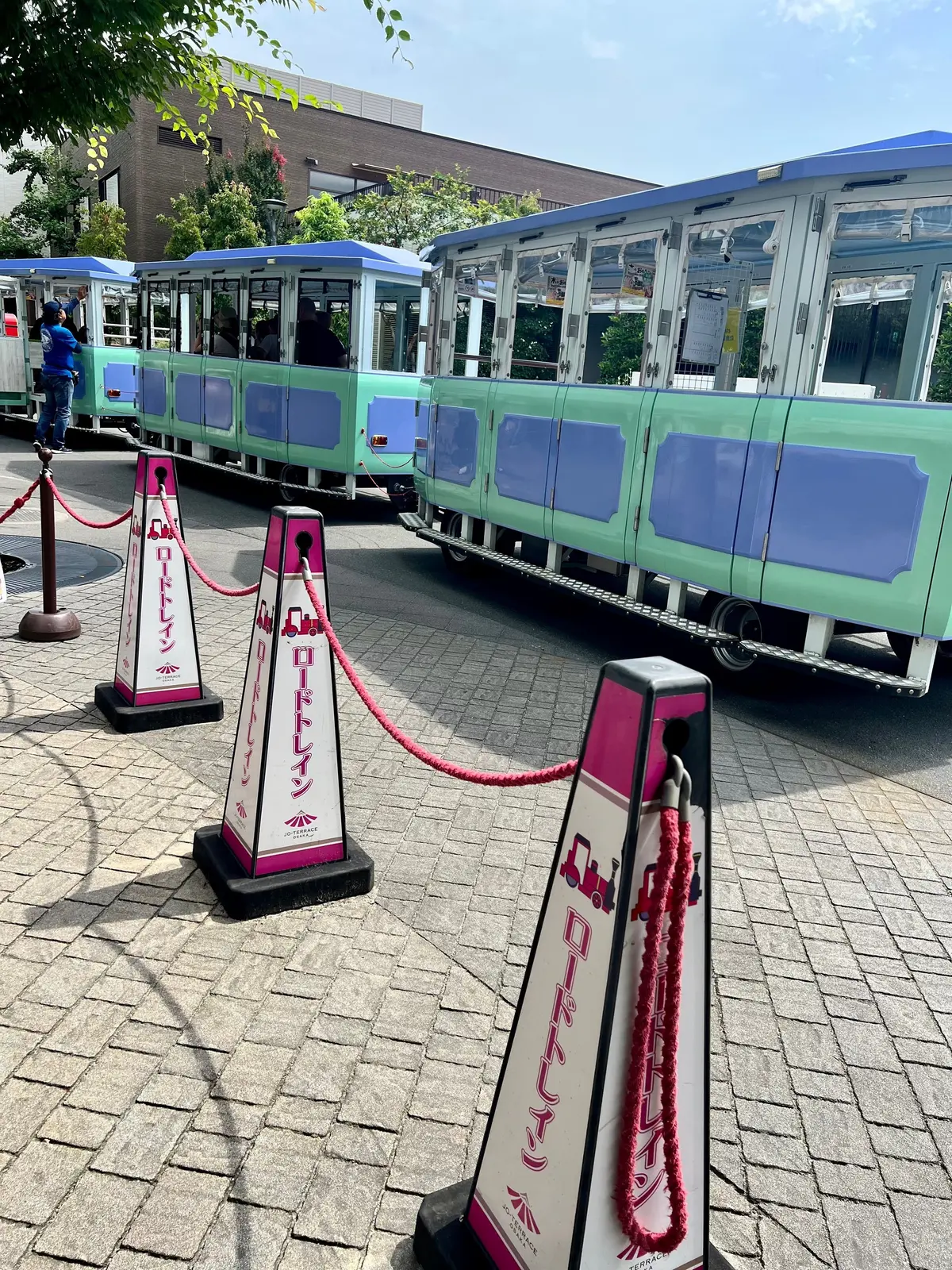 テラスゲートの駅構内。ライトグリーンのロードトレイン停車し、ピンクと黒のコーンで立ち入り禁止にされている。背景に建物と緑が見える。