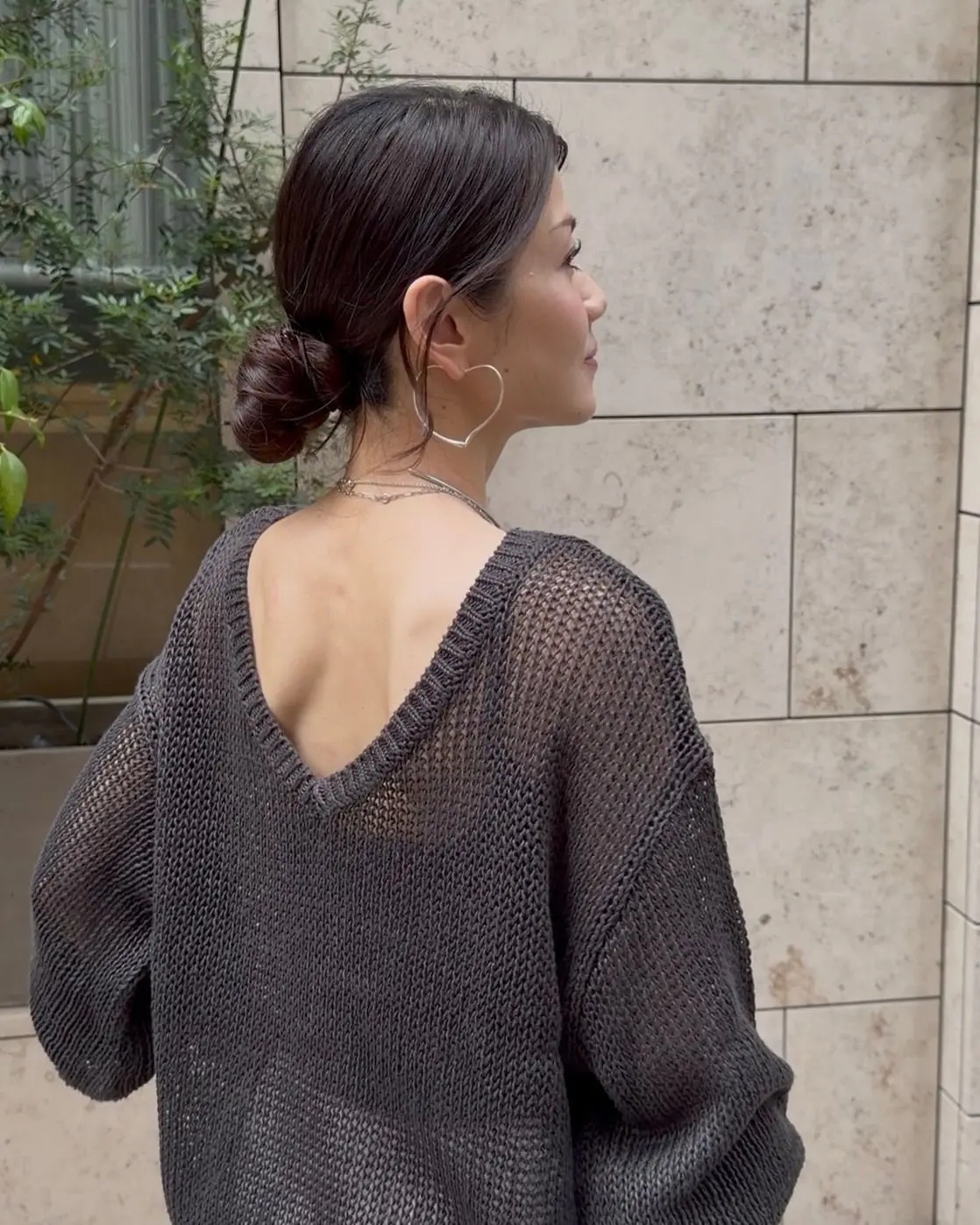 後ろから見た女性が、グレーのかぎ針編みのプルオーバーを着て、壁に寄りかかっている。耳にはハート型のイヤリングが輝いている。