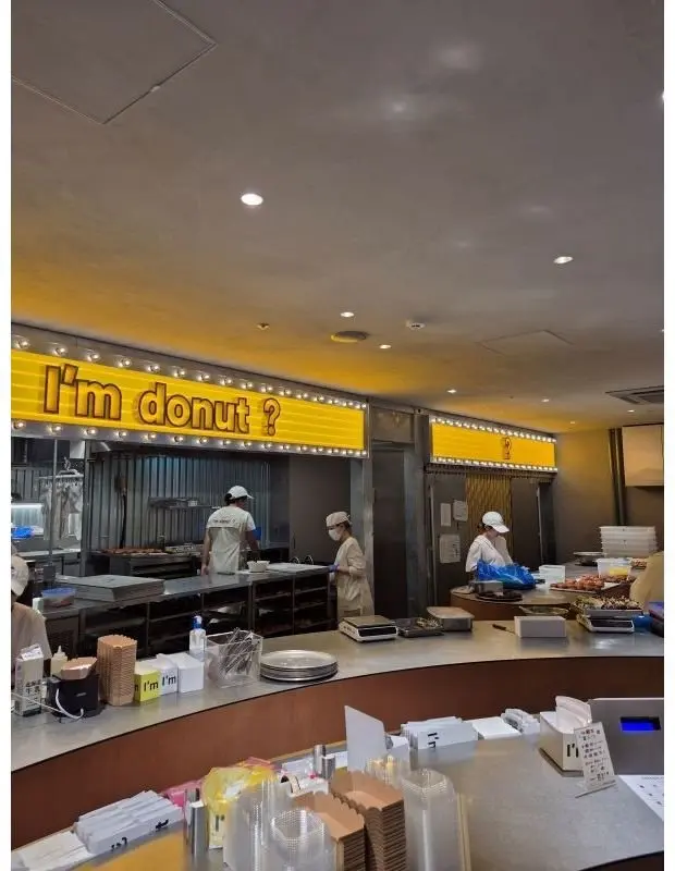 黄色い照明の店内で、イムド Donuts の看板が目立つ。カウンターには紙容器やトレイが並び、制服を着たスタッフがドーナツ作りや販売に勤しんでいる。