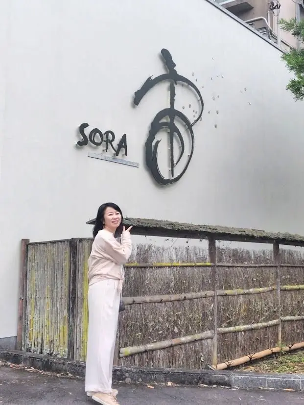 女性がベージュの服を着て、壁に「SORA」と書かれた看板の前でピースサインを作っている。壁は白で、看板は黒のデザイン。背景には竹製のフェンスと木製の屋根が見える。