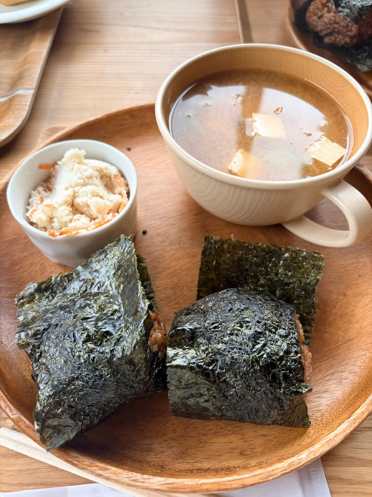 木製のプレートに、海苔で包まれたおにぎり2個、味噌汁、付け合わせの和え物が置かれている。おにぎりは三角形と四角形、味噌汁には豆腐が浮かんでいる。