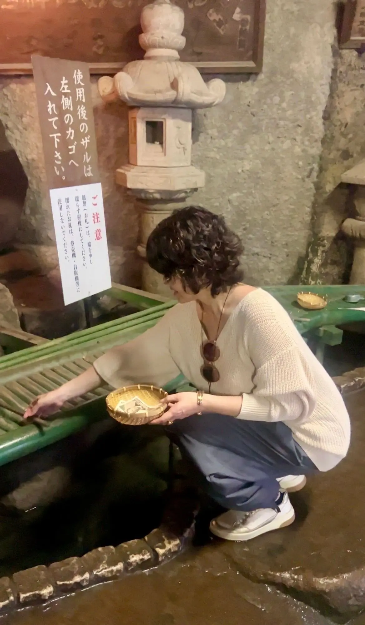 女性が石の噴水に手を伸ばし、小さな金色の器に水を汲んでいる。背後には石造りの灯篭と説明用の看板がある。