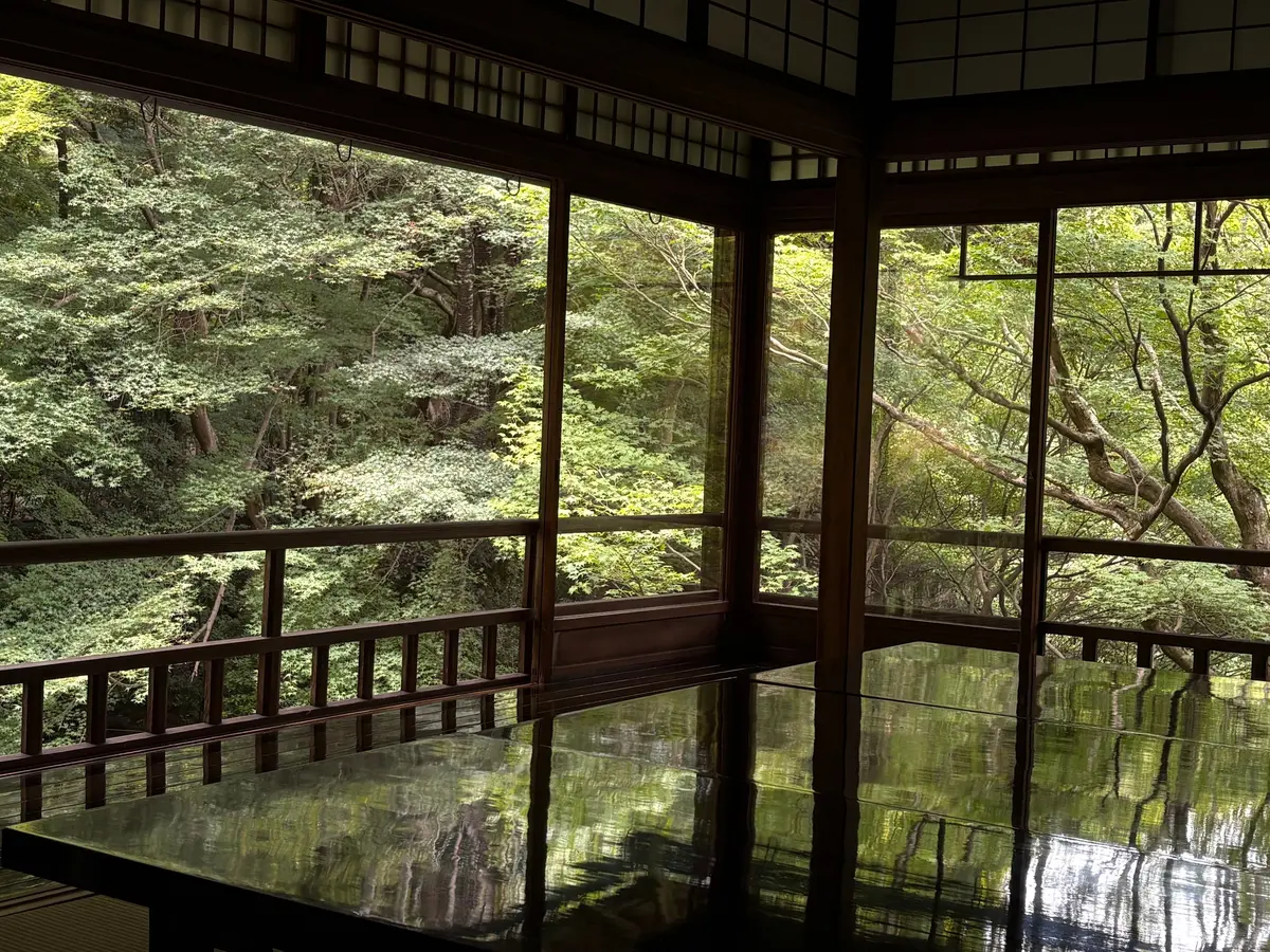 木々に囲まれた茶室の内部。木製の格子窓から緑豊かな庭園が望む。床は鏡のように光り、窓の外の景色を反射している。
