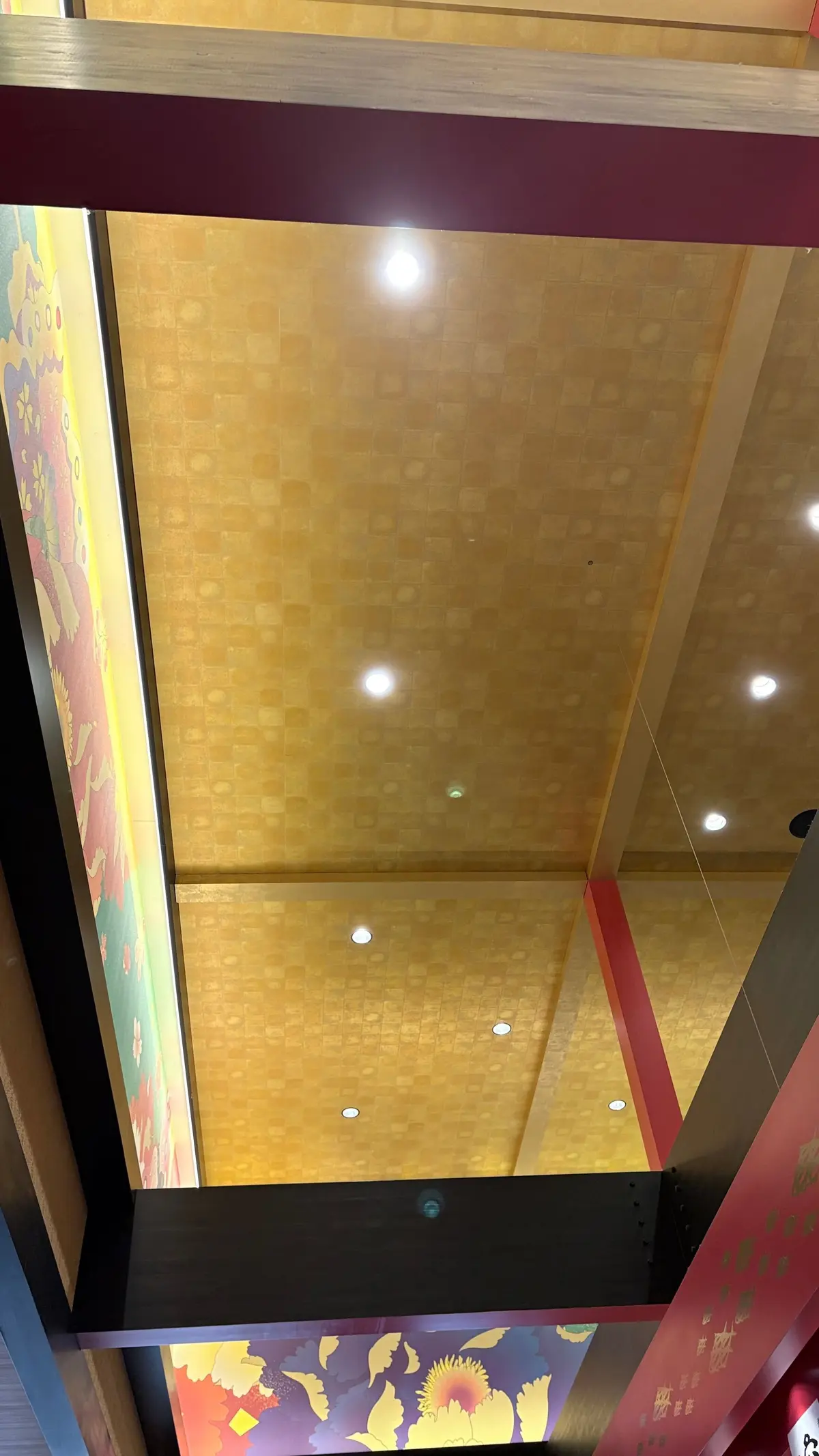 金色の幾何学模様の天井と、その下にあるカラフルな壁画。天井には白い照明が点灯し、壁画は花や人物が描かれている。赤紫色の梁が天井と壁画を隔てている。