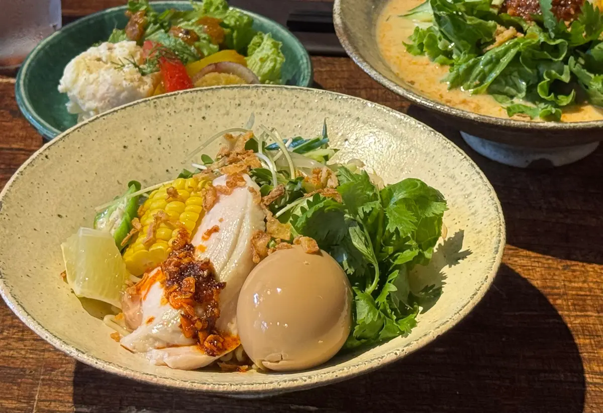 木製のテーブルに置かれた、黄色いそば猪口に入ったラーメン。麺の上に、茹で卵、トッピングされたコーン、パクチー、そして赤い辛味調味料が乗っている。背景には、他の料理が映る。
