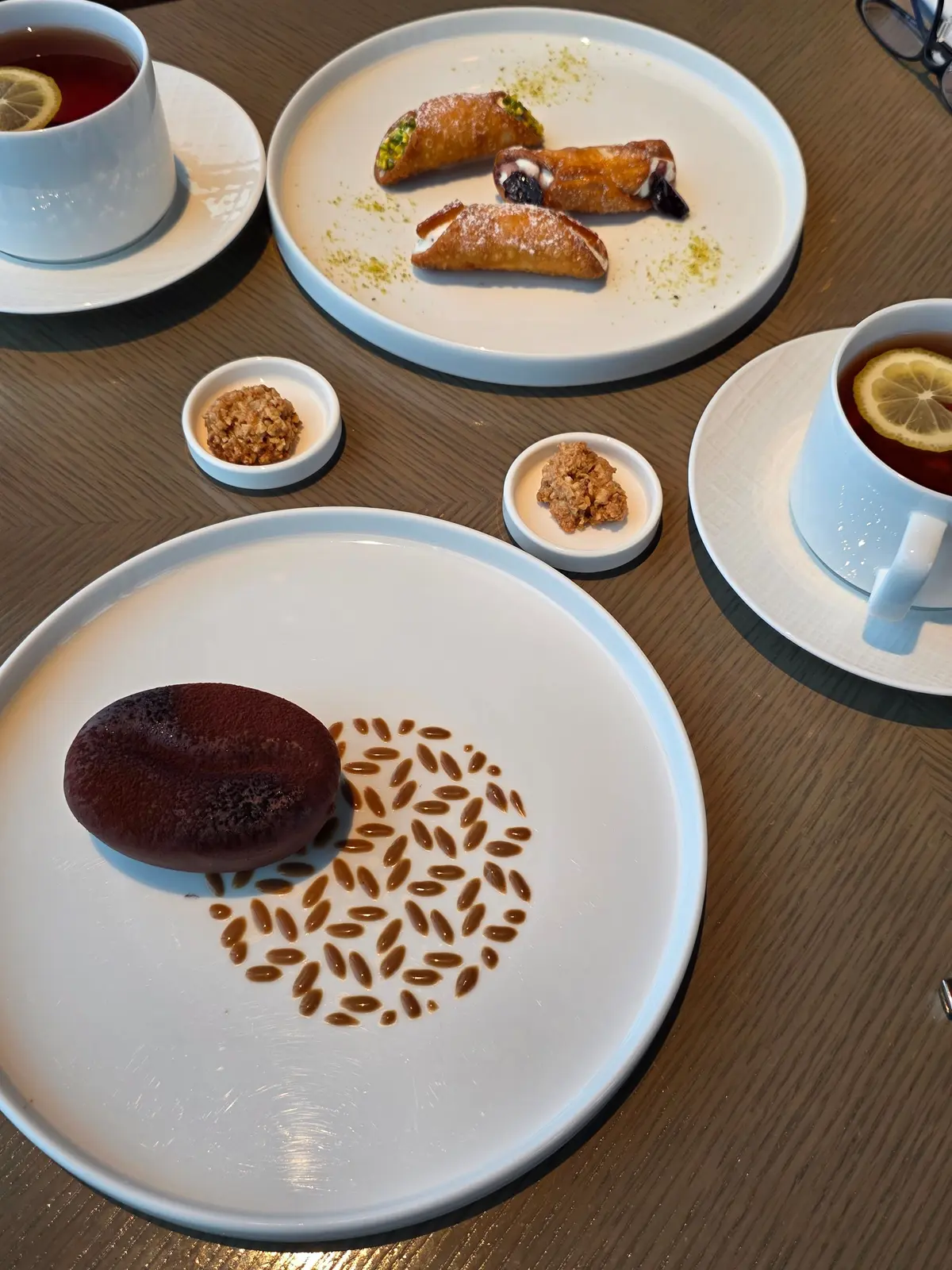 白テーブルに置かれた、トルコ菓子と紅茶の盛り合わせ。中央にチョコレートの塊と模様、上部にはカヌレのような菓子、右下にはレモン入りの紅茶。小皿にはナッツ菓子が添えられている。