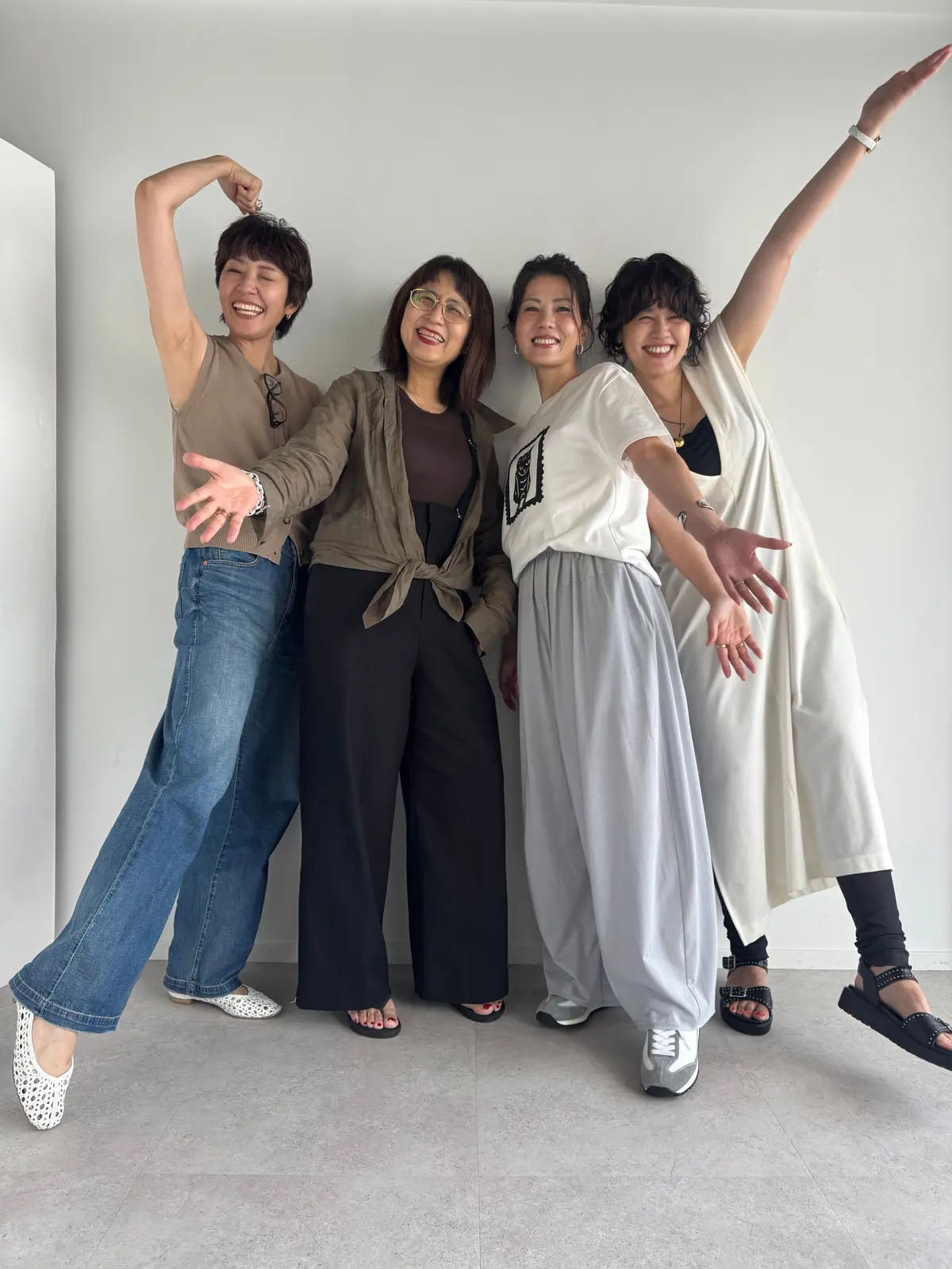 白い壁の前で、4人の女性が笑顔でポーズをとっている。左から、ベージュのタンクトップにデニム、ブラウンのカーディガンに黒のパンツ、白地に黒の模様のTシャツにグレーのパンツ、白いシャツワンピースを着ている。それぞれ腕を上げ、楽しげな雰囲気が伝わる。