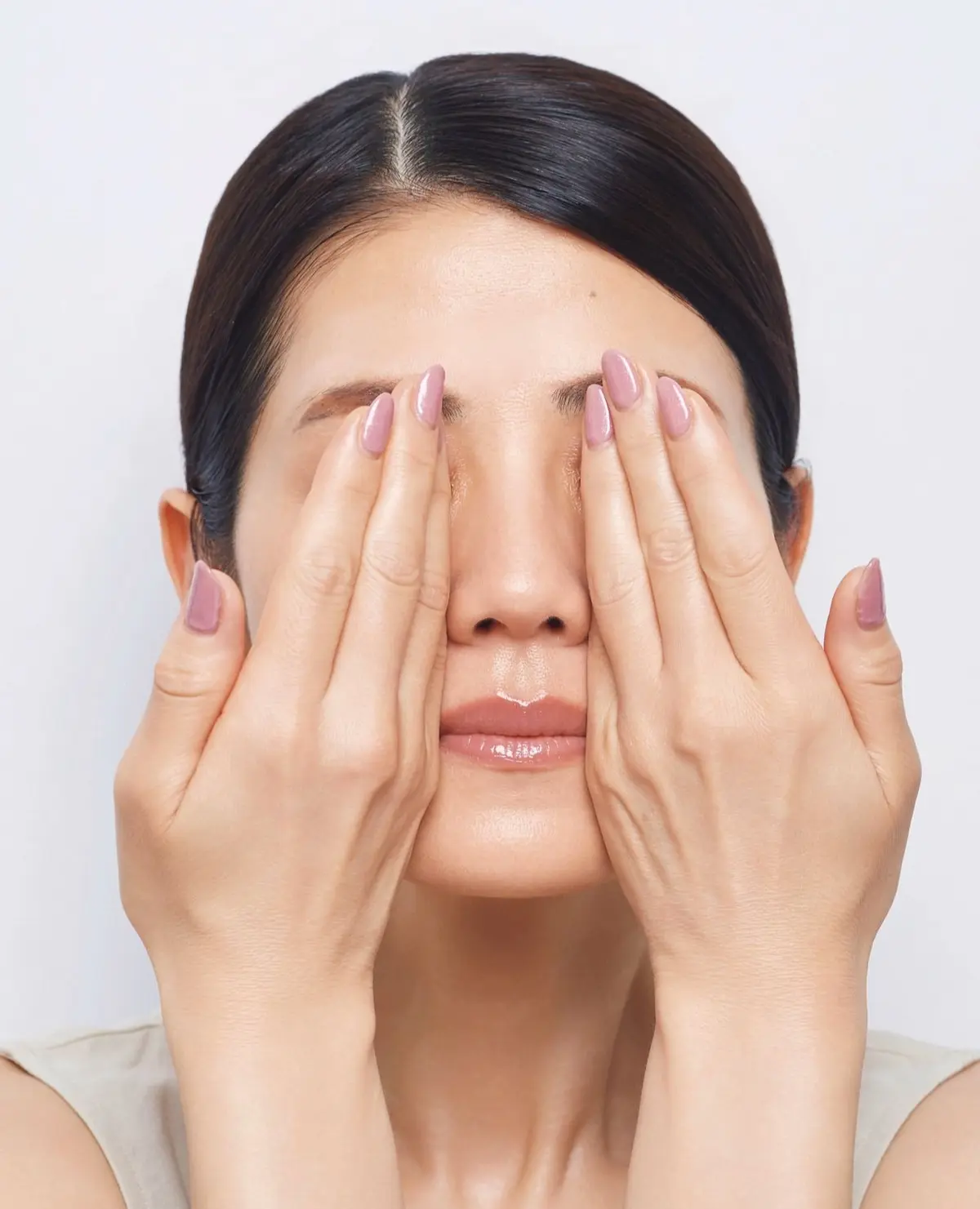 茶髪の女性が両手で目を覆い、正面を向いているポートレート。淡いベージュのトップスを着用し、ピンク色のネイルをしている。背景は白。
