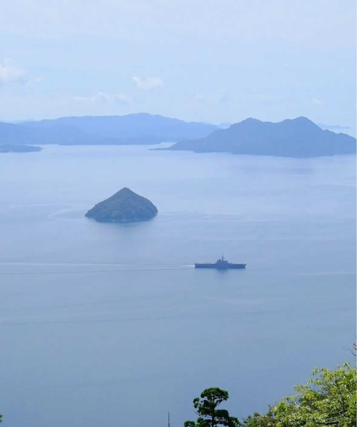 水平線上に、海に浮かぶ軍艦と小さな島、そして遠くの山々が連なる風景。手前には緑豊かな木々が生い茂っている。全体的に青みがかった、穏やかな雰囲気。