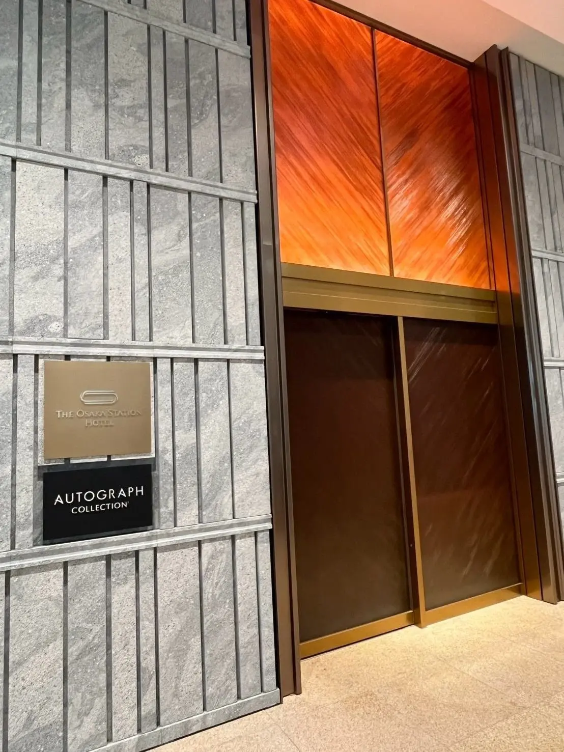グレーのタイル壁に設置された、The Osmosis Station Hotelの案内板とエレベーター。エレベーターのドアは閉まっており、壁面は木目調のオレンジ色。