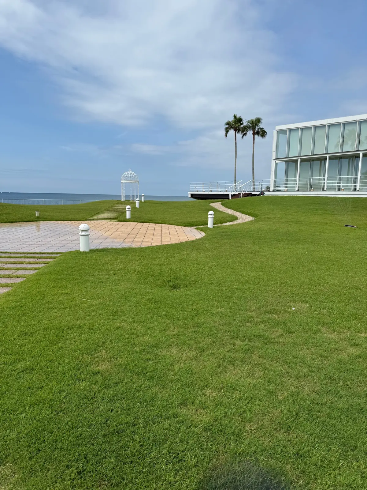 海辺の芝生に、白い欄干とベンチ、そして白い建物が見える風景。遠くには水面が広がり、青空に白い雲が浮かんでいる。