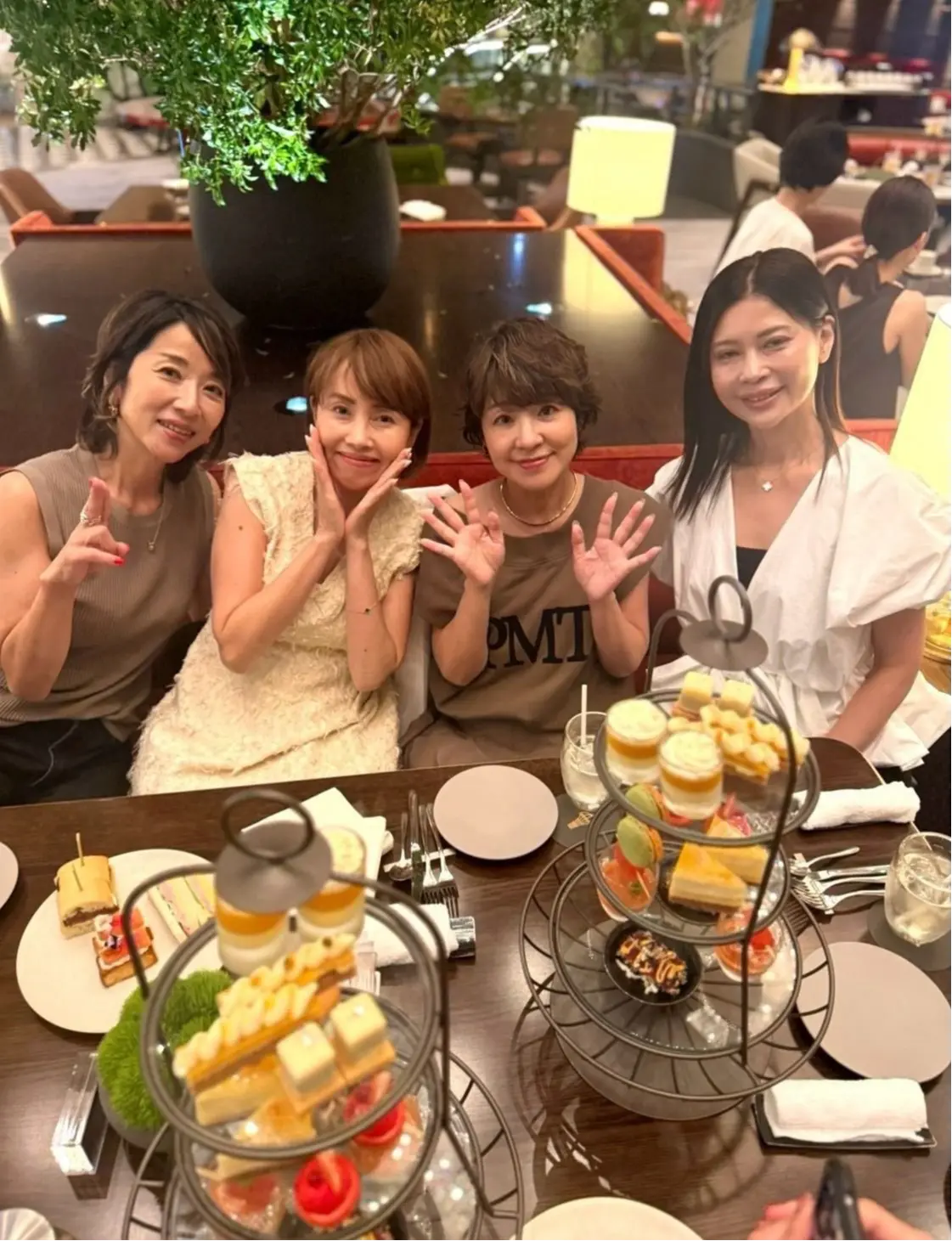 テーブルを囲み、笑顔でポーズをとる4人の女性。テーブルには三段重ねのスタンドに軽食が並び、グラスや食器も置かれている。背景にはレストランの様子が見える。