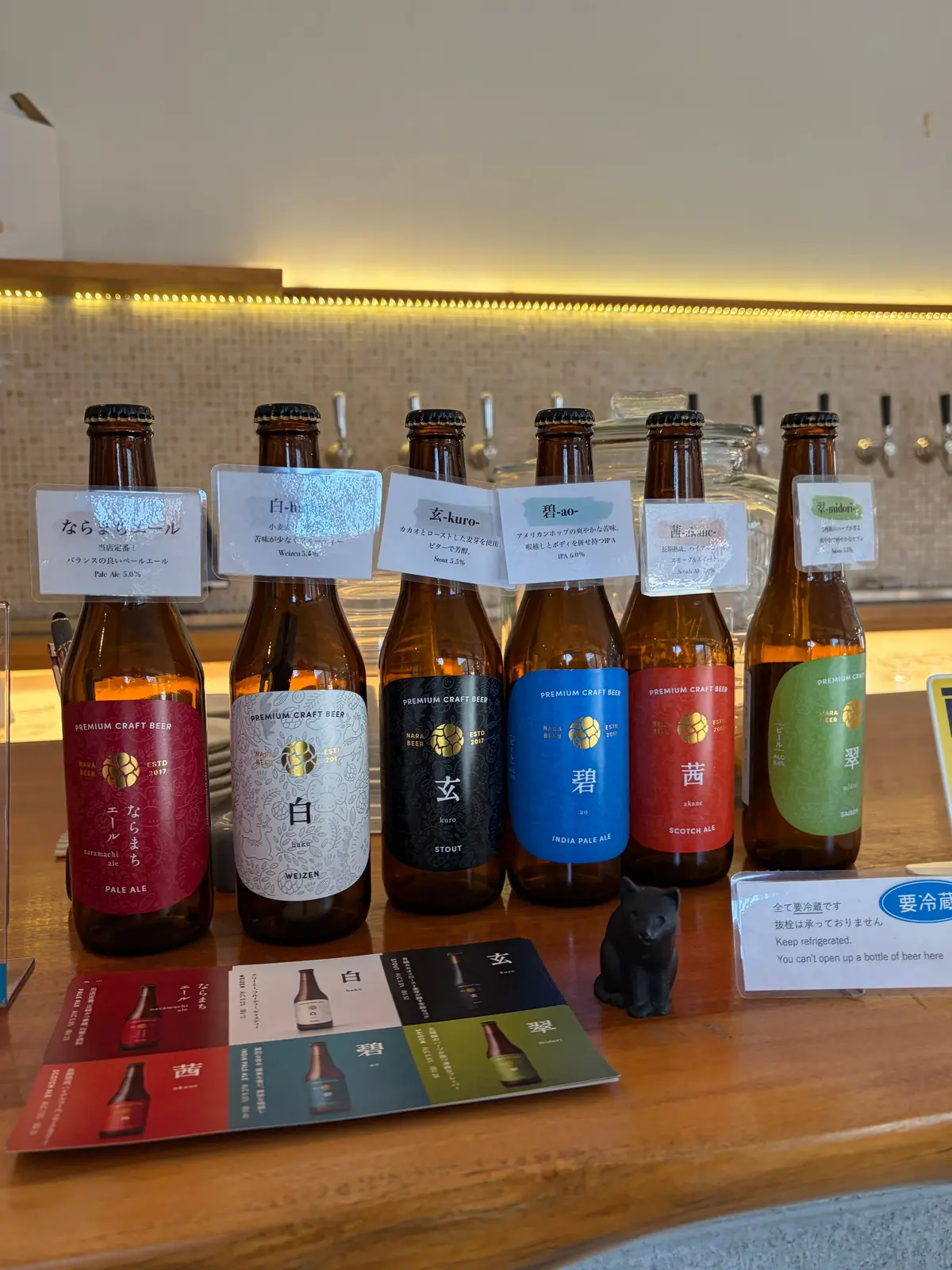 木製の台に並んだ、複数のクラフトビールの瓶。瓶には日本語のラベルが貼られ、それぞれ異なる種類のビール（ペールエール、白ビール、スタウト、インディアペールエール、スコッチエール、黒ビール）が示されている。瓶の前に黒い犬の置物がある。