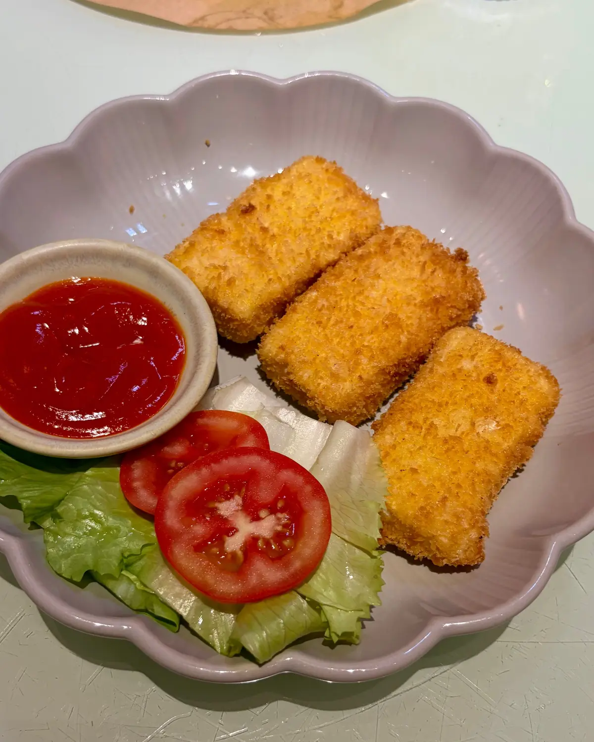 ピンク色の波状の皿に、四角いカツレツが3切れ、トマトのスライスとレタスの葉、赤いソースが入った小さなボウルが添えられている。