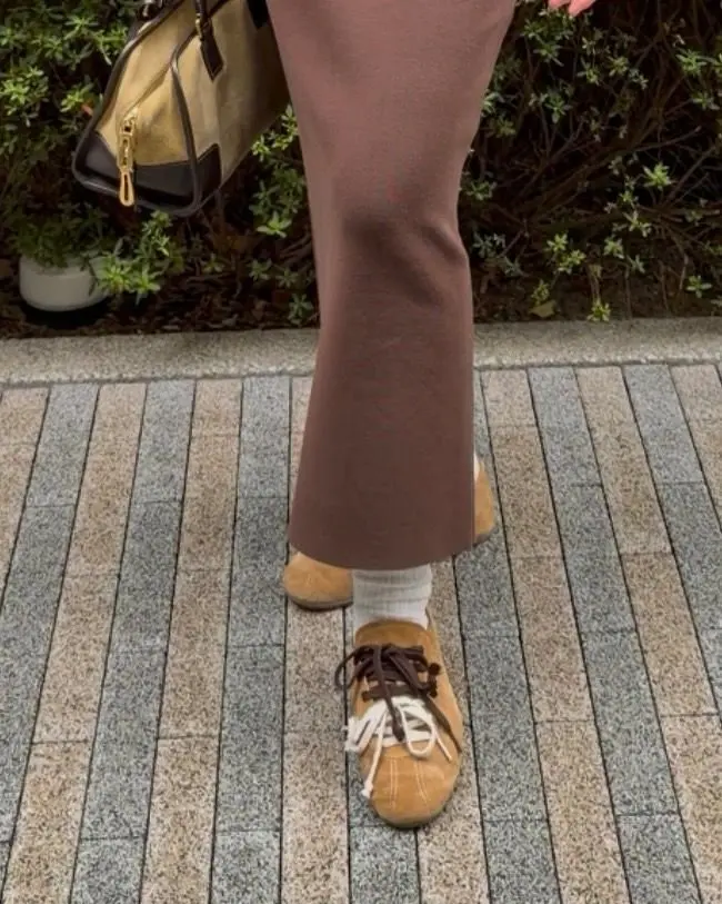 ブラウンのニットパンツとベージュのスエードローファーを履いた足元。白い靴下が見え、ローファーには茶色の紐が通っている。背景には金色のバッグと植物がある。
