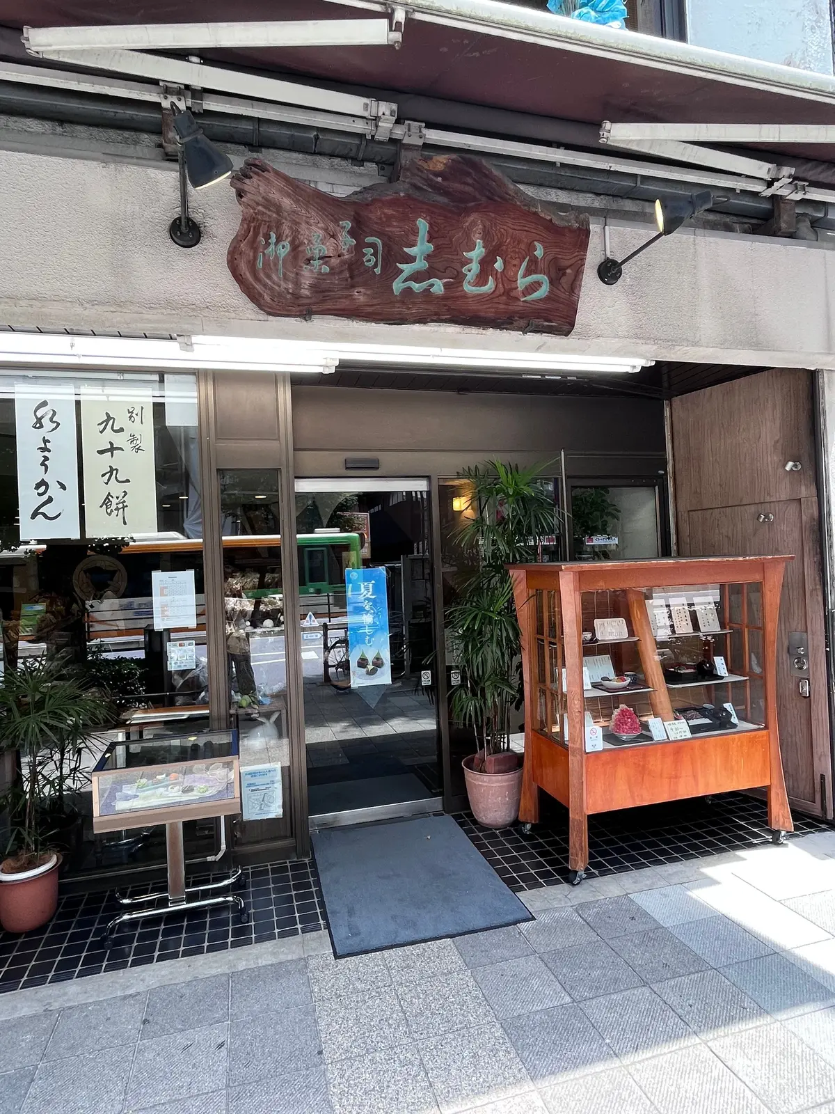 木造の建物にある「森むら」と書かれた店。入り口にはガラスケースがあり、商品が並んでいる。左には手書きの看板、右には木製のショーケースがある。植物が置かれ、落ち着いた雰囲気。