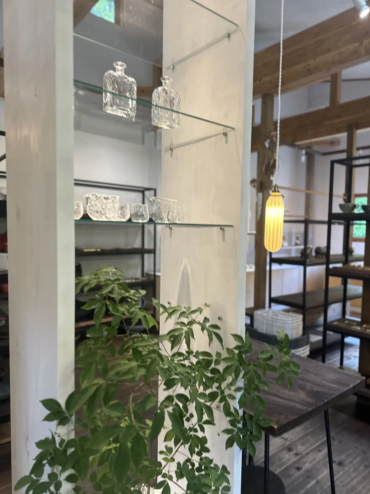ガラスの棚に透明な瓶が並び、温かい光のペンダントランプが下がる店内。木製のテーブルと緑の植物が配置され、モダンなインテリアが特徴。
