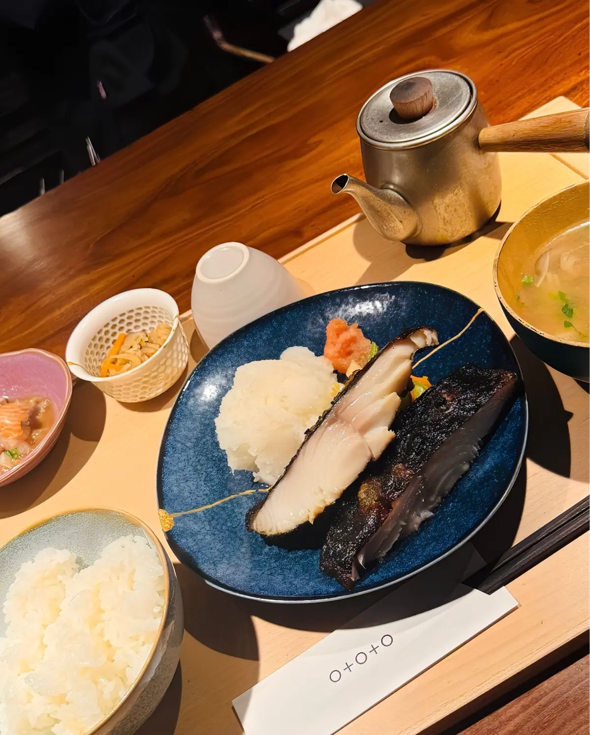木製のテーブルに置かれた和食の盛り合わせ。青い磁器皿に白米、魚の切り身、ネギ、赤身の魚の部位が乗っている。茶碗に入った白米、小鉢に入ったピンク色の料理、黄色い汁物、お茶が入った急須が添えられている。箸と紙ナプキンも写っている。