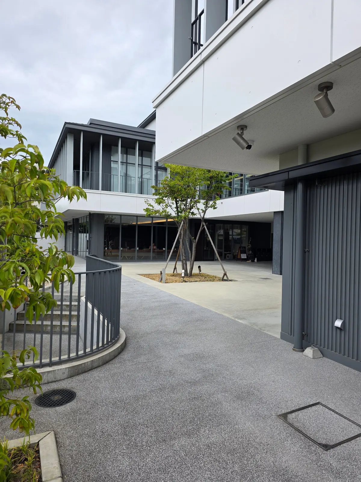 灰色の舗装された通路が、白い現代的な建物群の間を縫うように伸びている。通路沿いには手すりが設置され、中央には若い木が植えられている。背景には、窓や入り口のある建物が並び、全体的にシンプルなデザインが特徴。