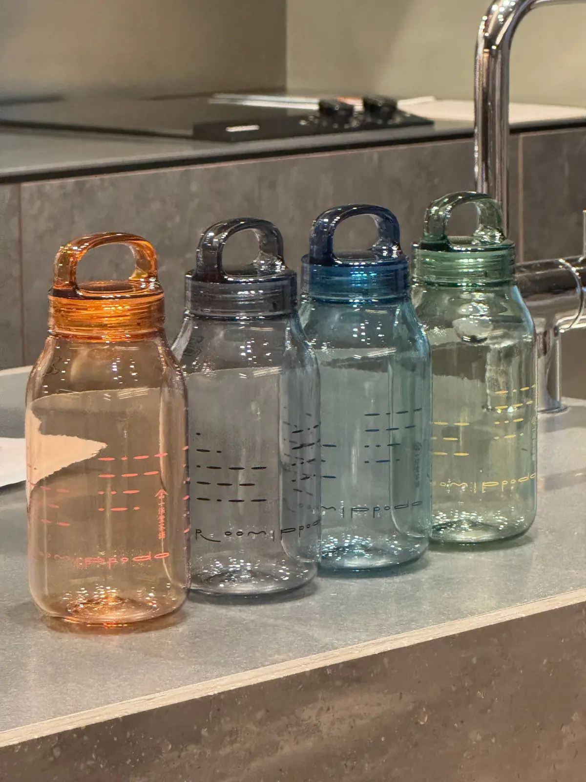 キッチンカウンターに並んだ4つの水筒。左からオレンジ、黒、青、緑のフタ付きで、それぞれ異なる色とデザイン。