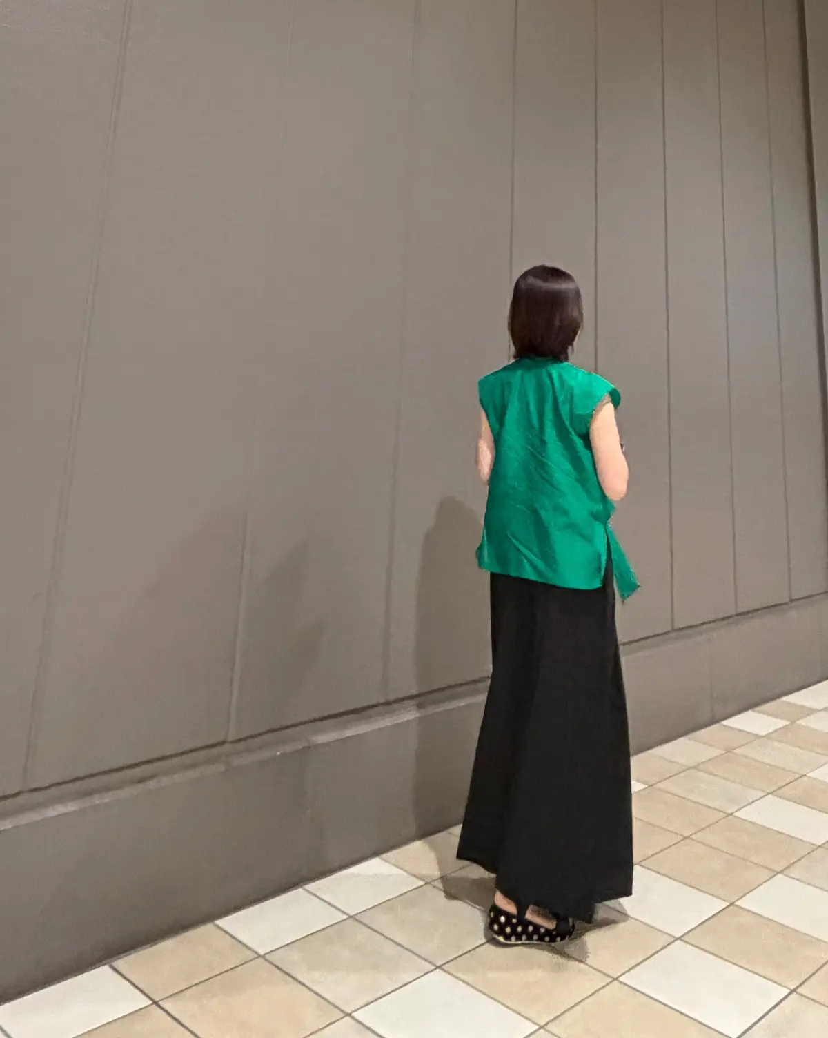 黒いロングスカートと緑色のトップスを着た女性が、グレーの壁の前に立っている。女性は後ろ姿で、タイルの床に足を踏み入れている。