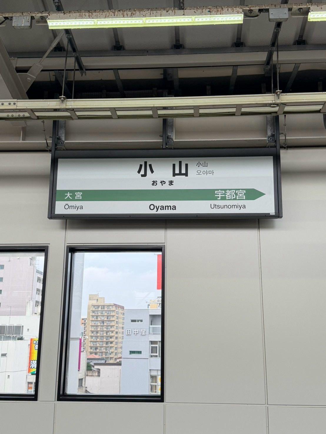 鉄道駅の標識。駅名「小山（おやま）」が日本語、韓国語、ローマ字で表示。左右に「大宮」「宇都宮」と駅名が示されている。背景には窓から見える高層ビル群。