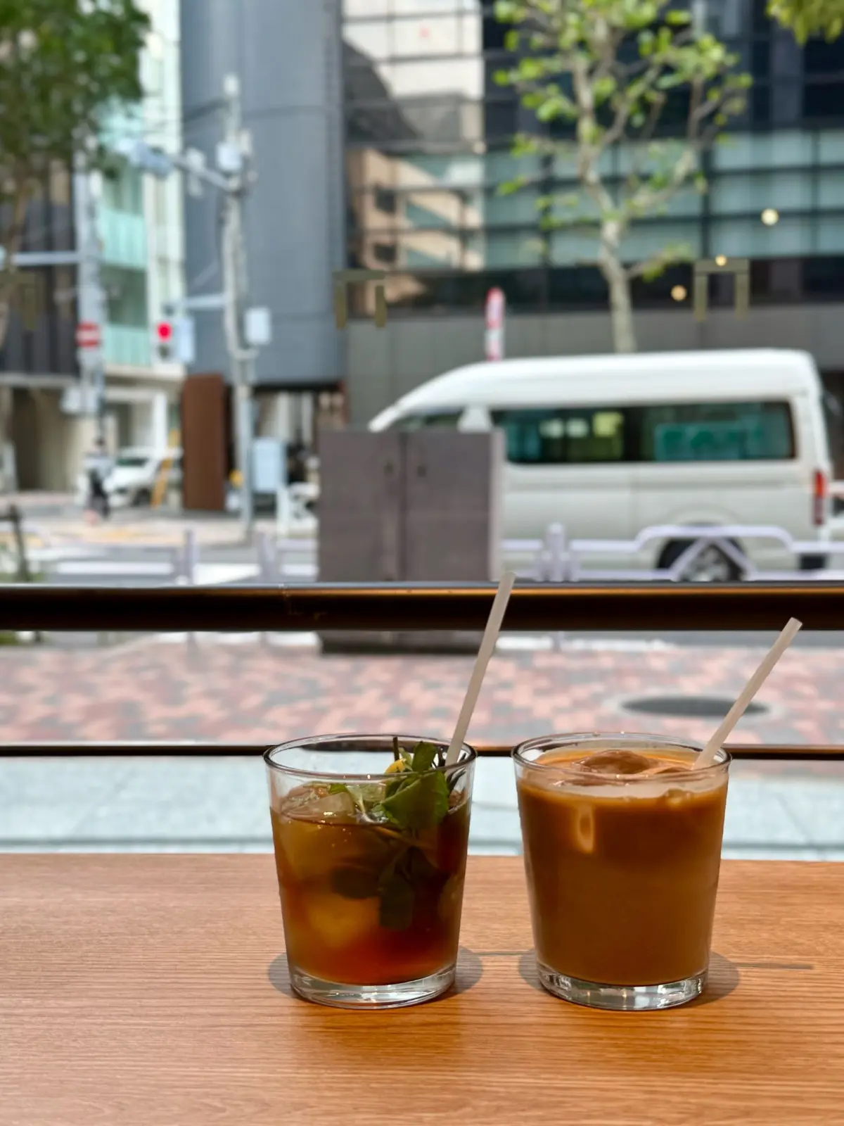 木製のテーブルに置かれた2つのグラス。左は柑橘系の入ったお茶、右はコーヒー。それぞれにストローが挿さっており、背景には都市の風景が見える。