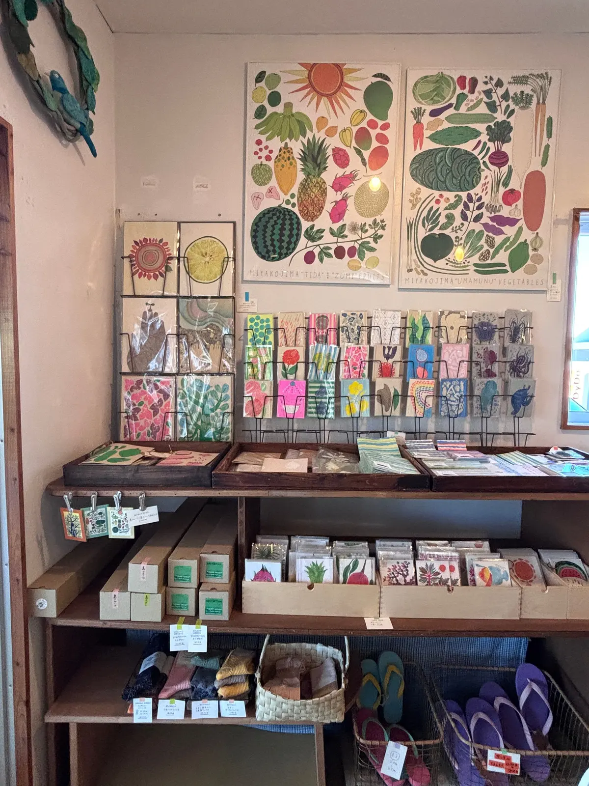 カラフルなイラストの壁掛けが飾られたお店のディスプレイ。棚には、野菜や果物の絵柄のカード、ノート、石鹸などが並び、手作りのような温かみのある雰囲気が感じられる。