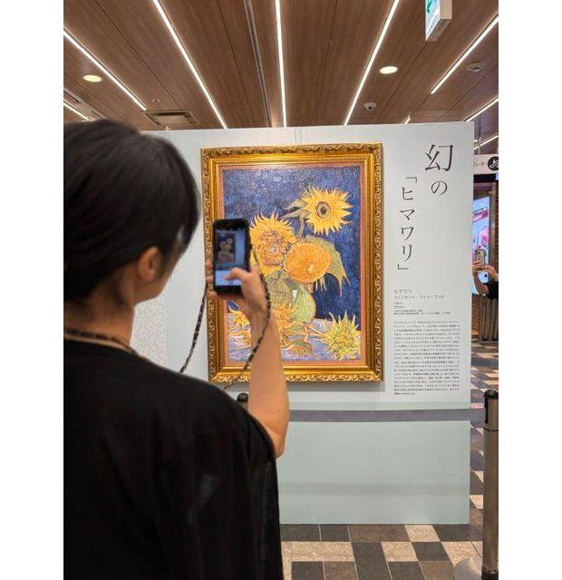 黒髪の女性がスマートフォンで、金縁の額縁に入ったひまわり絵画を撮影している。背景には日本語の解説板と、商業施設の様子が見える。