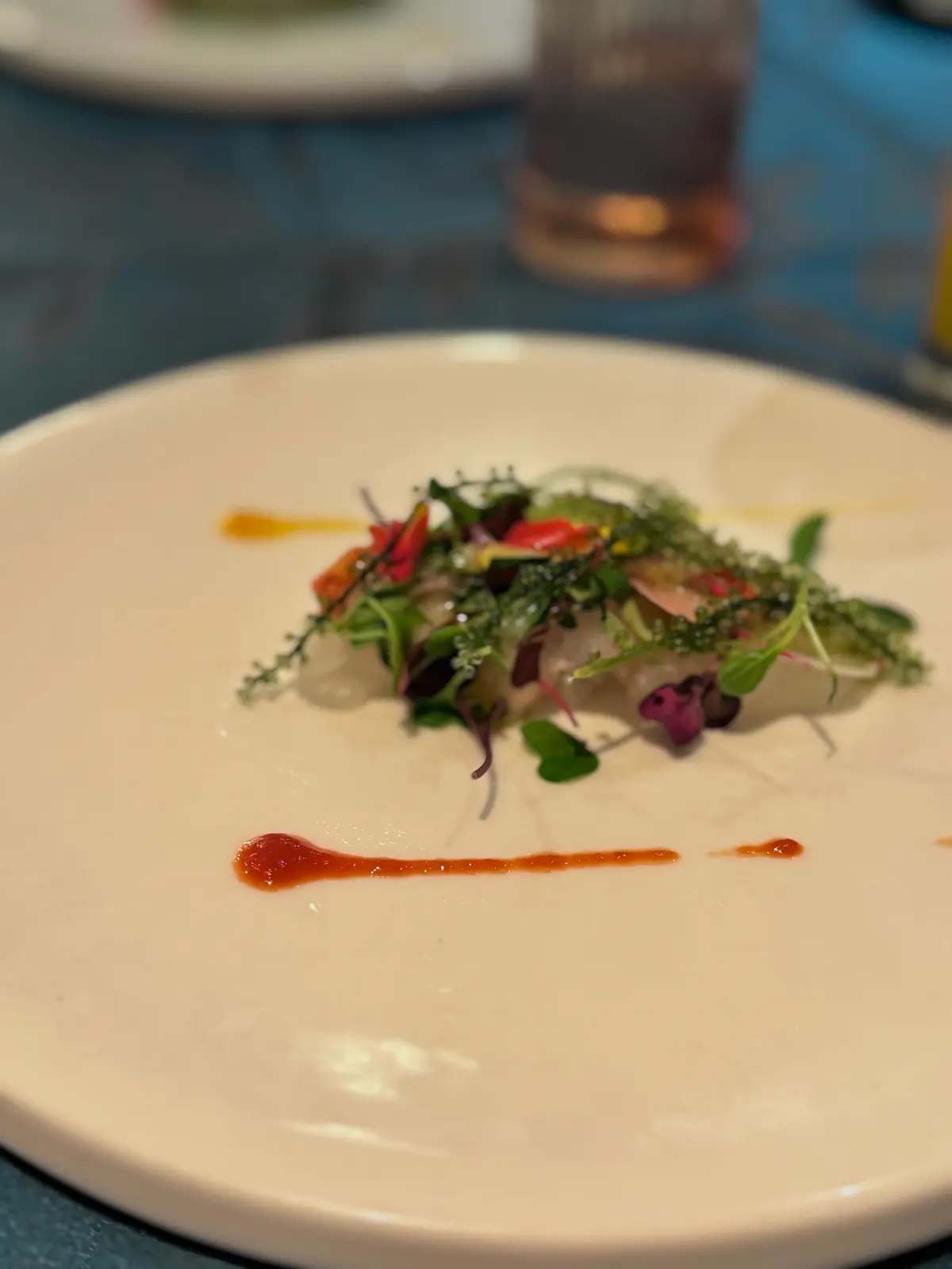 白磁の皿に盛り付けられた、魚介を使った料理。魚の周りには緑色の海藻や、赤や緑の小さな葉野菜が彩りを添えている。皿の下には、赤とオレンジ色のソースが線状に添えられている。