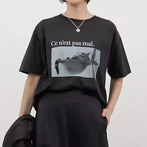 黒いTシャツに、猫の画像とフランス語のテキスト「Ce n'est pas mal.」がプリントされている。Tシャツはゆったりとしたシルエットで、モデルが着用している。