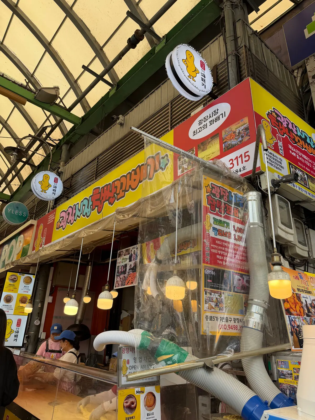 カラフルな看板が並ぶ屋台街の店先で、黄色の看板と赤い看板が目立つ。店内には数人の客が並び、照明が明るく店内を照らしている。