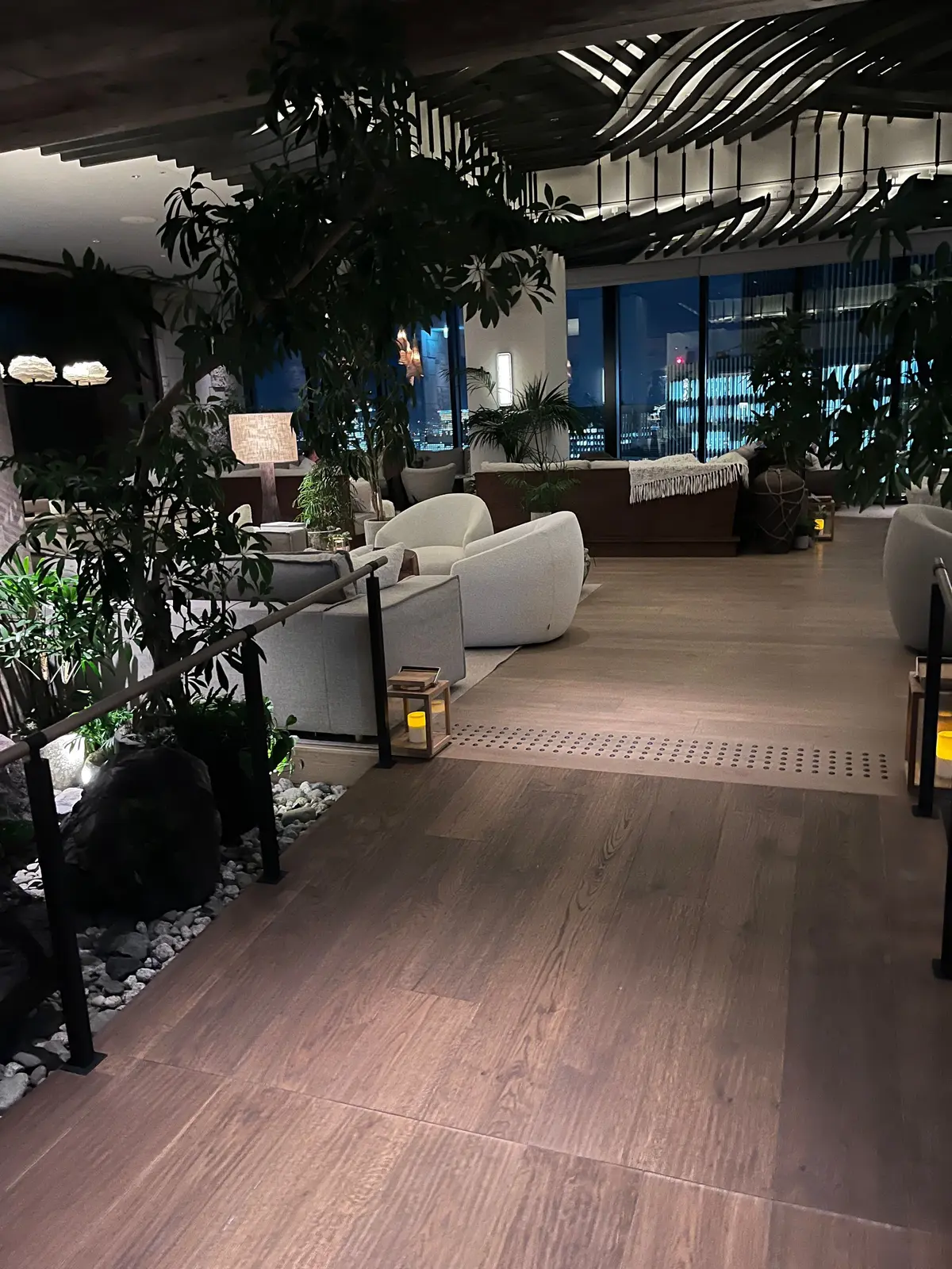 夜景を望む、モダンな内装のラウンジ。木目調の床に、白いソファと観葉植物が配置され、リラックスできる空間を演出。手すりや石積み、ランタンがアクセントになっている。