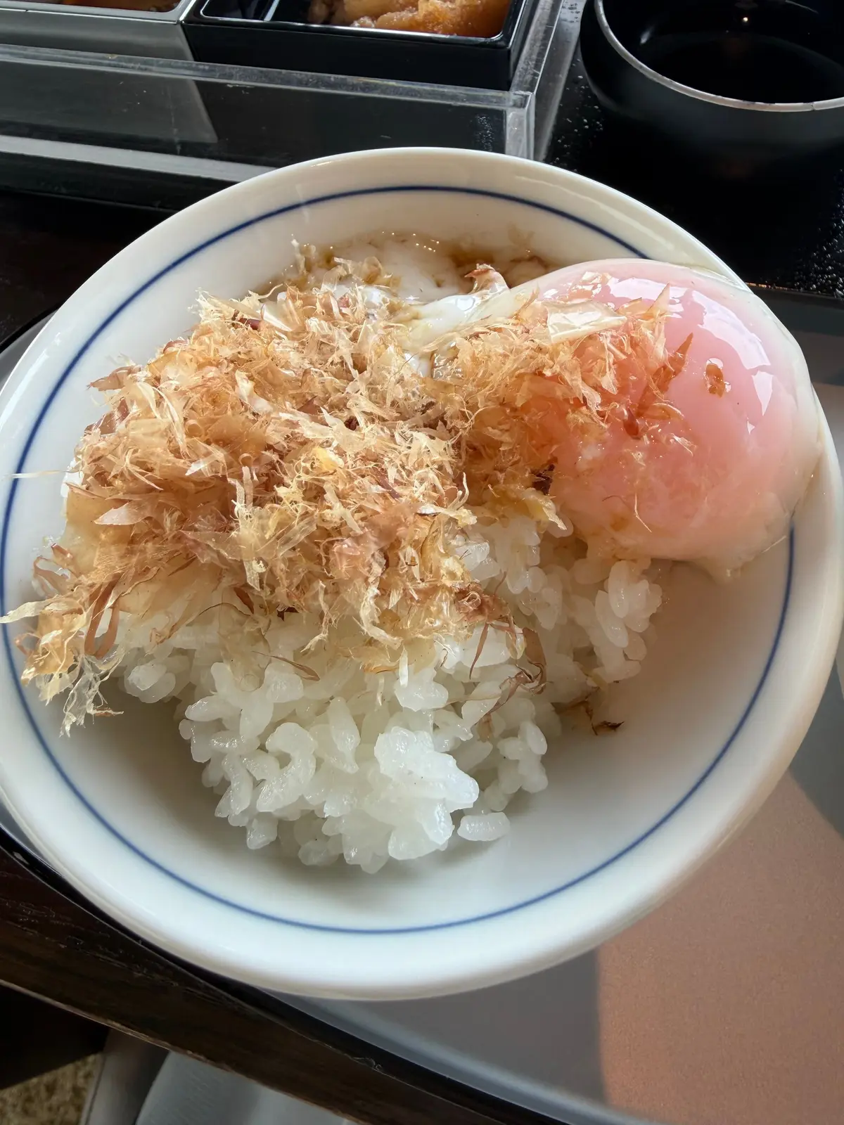 白くて丸い卵の上に、茶色いふわふわのかつお節が乗ったご飯茶碗。白い縁取りのある陶器の器に入っている。