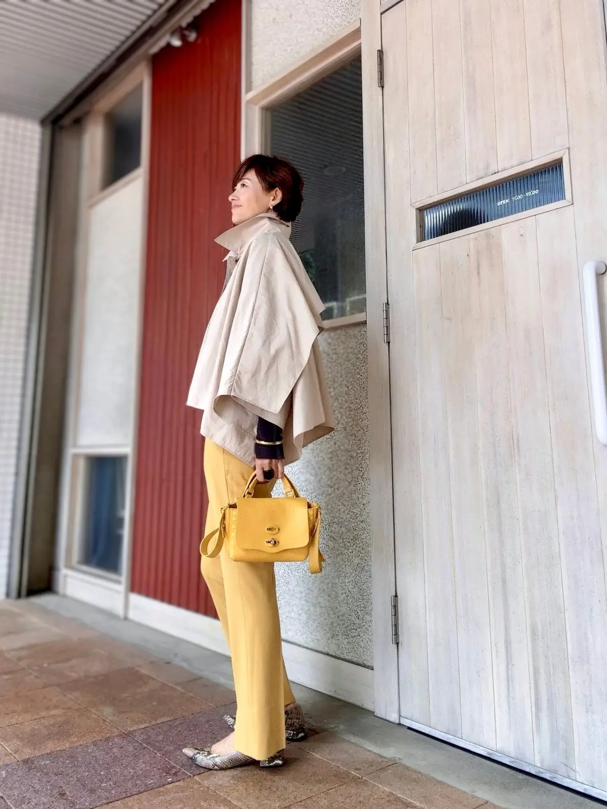 ベージュのケープに黄色いパンツ、黄色いハンドバッグを合わせた女性。茶色のショートヘアで、建物の前で横を向いている。