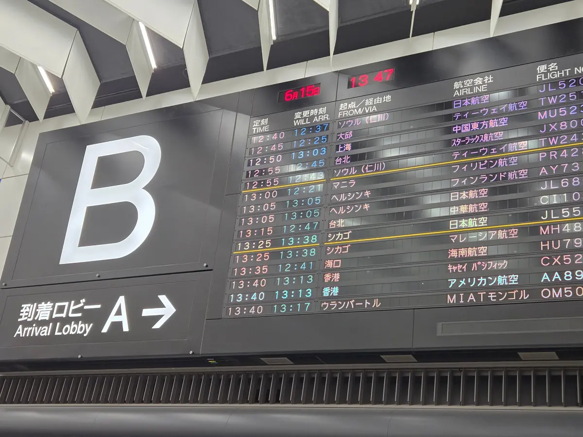 空港のフライト情報表示板。6月15日13時47分時点の国内線出発便情報が緑色で表示され、右側に航空会社名が列挙されている。左下には「到着ロビー A →」の案内表示。