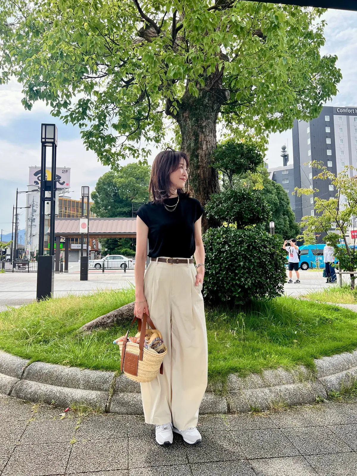 黒いカットソーにベージュのカーブパンツ、白いスニーカーを合わせた。ロエベのカゴバッグ、木陰に立っている。背景には街並みと高層ビルが見える。
