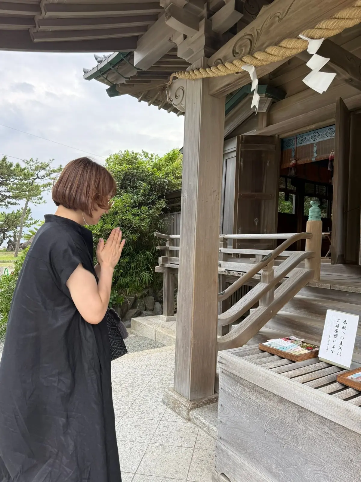 木造の神社で、女性が手を合わせ祈りを捧げている様子。黒いワンピースを着ており、背景には緑の木々が見える。神社の屋根には飾りがあり、入り口には注連縄が飾られている。